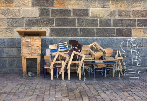 A collection of discarded wooden chairs and stools, some stacked or leaning against each other, arranged on a paved outdoor surface in front of a grey brick wall. The chairs are made of light and dark wood with different finishes, and some have metal or plastic components. To the left, there is a tall, vertical wooden cabinet or storage box with a lid partially open, positioned directly against the wall. On the right, a metal trolley or frame with a circular top and handle is visible. The overall scene depicts outdoor debris likely awaiting rubbish removal, with the setting suggesting a back alley or service area in Victoria. The lighting is natural, casting soft shadows on the cobblestone ground, fitting a typical rubbish clearance context for Commercial Waste Victoria.