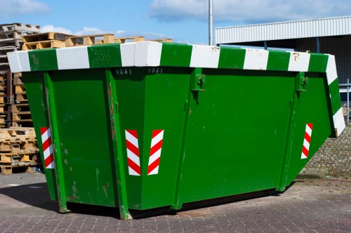 A large green industrial rubbish skip situated on a paved driveway, used for waste removal services by Commercial Waste Victoria. The skip has a rectangular shape with high sidewalls, constructed from durable metal with a smooth, painted surface, and features horizontal white and green reflective safety stripes on the sides. The front of the skip demonstrates a large, hinged metal door with a small latch for secure sealing. In the background, there are stacks of wooden pallets, and part of a warehouse or commercial building with a metal wall is visible, indicating an industrial or commercial area in or near Victoria. The lighting suggests an overcast day, giving soft natural illumination to the scene. This image illustrates the typical equipment used in waste management operations involving rubbish collection and disposal, aligning with services offered by Commercial Waste Victoria in the local area.