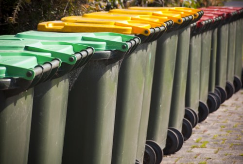 A row of large, cylindrical wheelie bins is lined up along a paved outdoor area, with three distinct colour-coded lids in green, yellow, and red, indicating different waste categories. The bins are made of durable plastic with a smooth finish and are positioned closely together, their black wheels resting on the pavement. The background features some greenery and a hedge, suggesting a residential or commercial setting within Victoria. The lids are securely closed, and the bins are clean and well-maintained, reflecting professional waste management services typical of organisations like Commercial Waste Victoria. The scene portrays a clean, organized waste collection point on a daylight day, suitable for rubbish removal or waste disposal purposes in the local area.