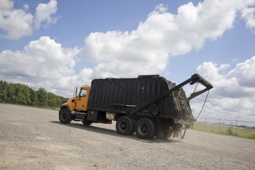 A large, yellow commercial waste collection truck with a black waste container on its back is parked on a gravel surface under a partly cloudy sky. The container is open at the top, revealing dark waste materials inside, and a hydraulic lifting arm extends from the truck, tilted upward at an angle. The background features a rural landscape with green trees and open fields, suggesting an outdoor rubbish removal operation near Victoria. The scene is well-lit with natural daylight, emphasizing the sturdy metallic surfaces of the truck and the textured gravel ground, illustrating a typical waste management activity conducted by Commercial Waste Victoria in the region.