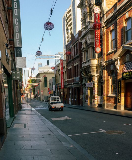 A narrow urban alleyway with a slight incline, lined with tall, closely packed buildings made of brick and concrete in muted tones of brown, gray, and red. On the right, a black building with metal sh