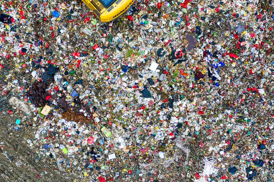 An aerial view of a large, uneven pile of mixed general waste composed of plastic bottles, food wrappers, cardboard pieces, clothing scraps, and other household refuse, scattered across an outdoor gro